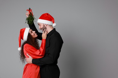 Photo of Lovely couple in Santa hats under mistletoe bunch on grey background. Space for text