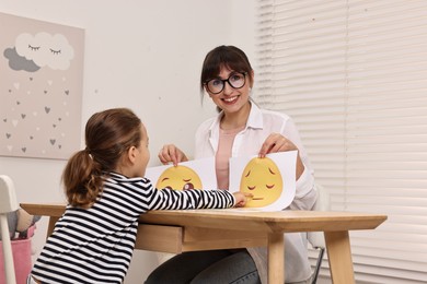 Photo of Autism therapy. Little girl choosing emoticon at table with smiling psychologist in mental health center