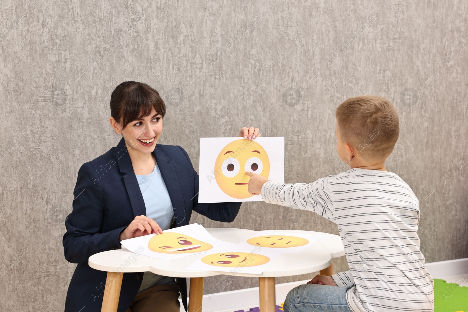 Photo of Autism therapy. Little boy choosing emoticon at table with smiling psychologist in mental health center, back view