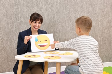 Photo of Autism therapy. Little boy choosing emoticon at table with smiling psychologist in mental health center, back view