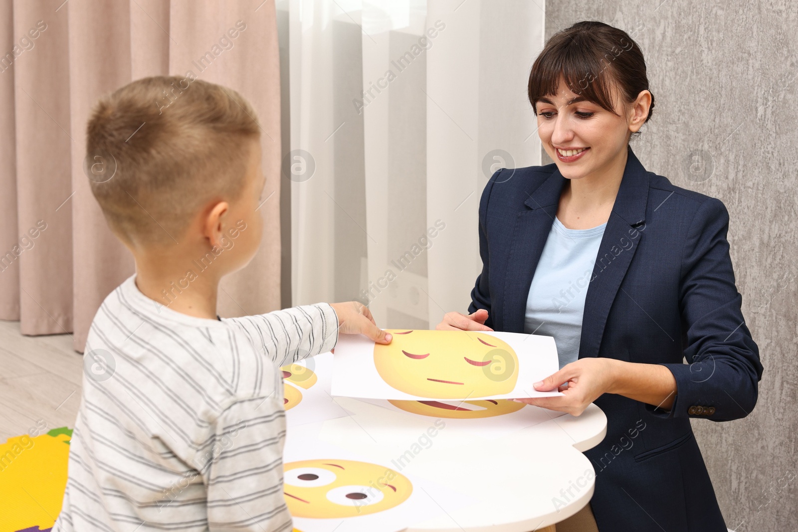 Photo of Autism therapy. Little boy choosing emoticon at table with smiling psychologist in mental health center, back view