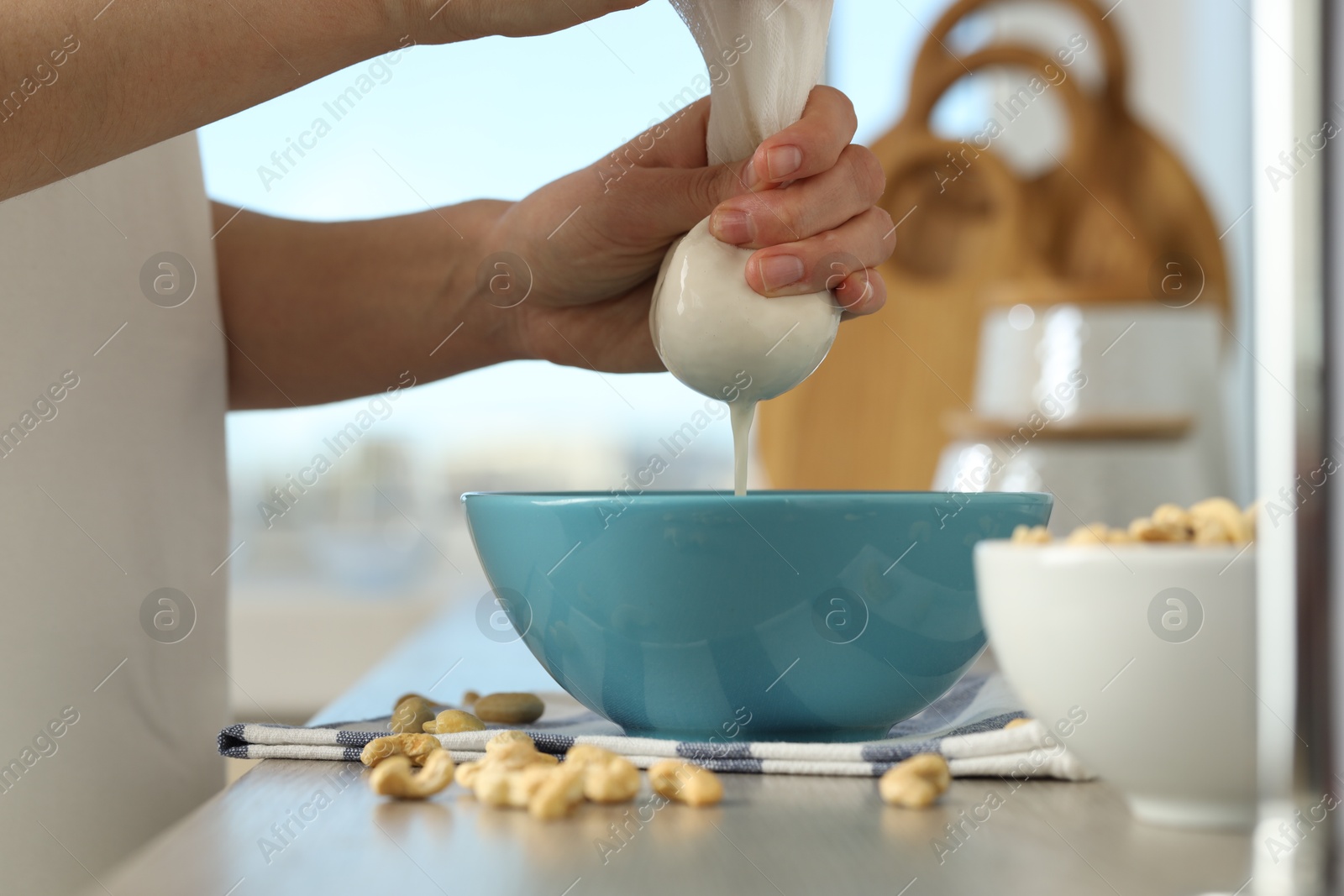 Photo of Woman straining cashew milk into bowl at light table with nuts indoors, closeup