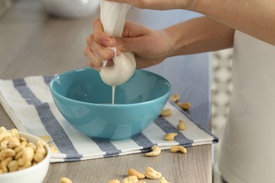 Woman straining cashew milk into bowl at wooden table with nuts indoors, closeup