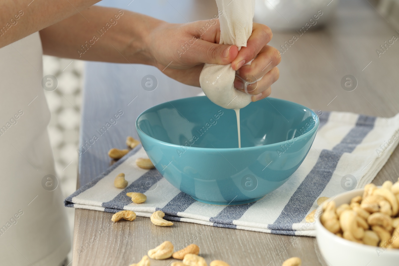 Photo of Woman straining cashew milk into bowl at wooden table with nuts indoors, closeup