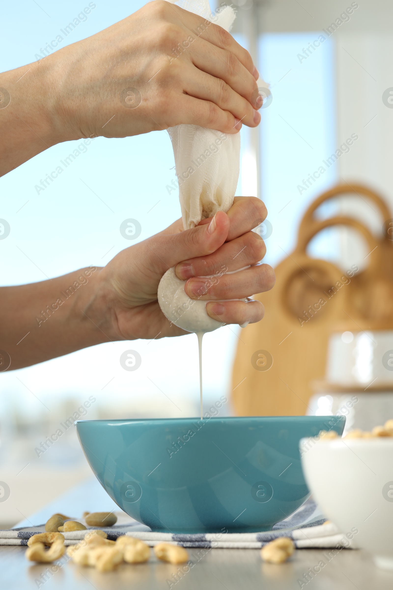 Photo of Woman straining cashew milk into bowl at white table with nuts indoors, closeup