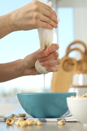 Woman straining cashew milk into bowl at white table with nuts indoors, closeup