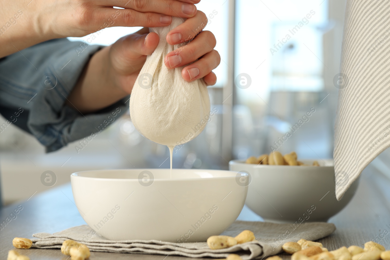 Photo of Woman straining cashew milk into bowl at wooden table with nuts indoors, closeup