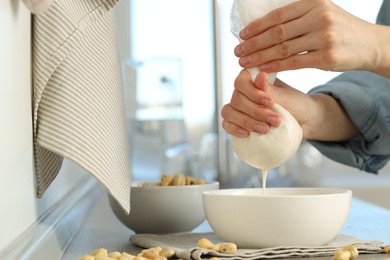 Woman straining cashew milk into bowl at wooden table with nuts indoors, closeup