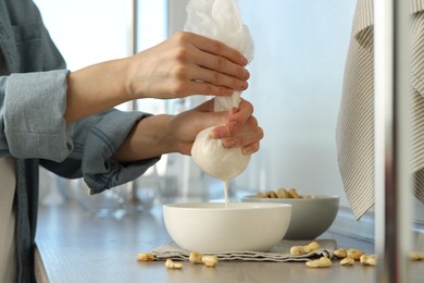 Photo of Woman straining cashew milk into bowl at wooden table with nuts indoors, closeup