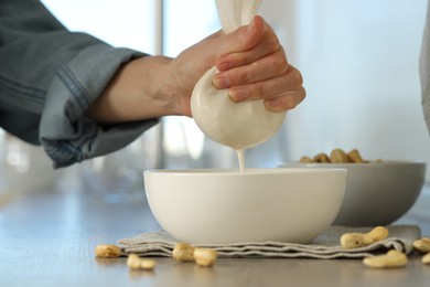 Photo of Woman straining cashew milk into bowl at wooden table with nuts indoors, closeup