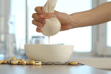 Woman straining cashew milk into bowl at light table with nuts indoors, closeup