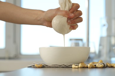 Woman straining cashew milk into bowl at light table with nuts indoors, closeup