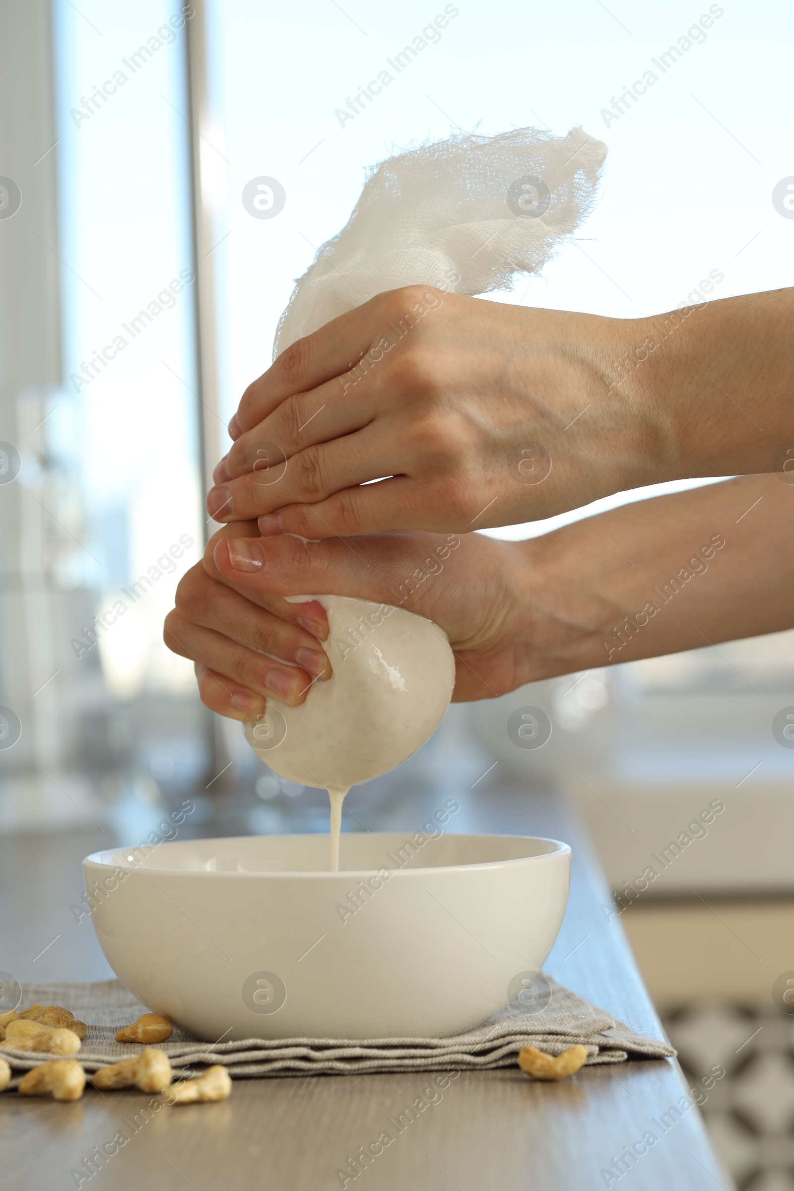 Photo of Woman straining cashew milk into bowl at wooden table with nuts indoors, closeup