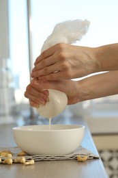 Photo of Woman straining cashew milk into bowl at wooden table with nuts indoors, closeup