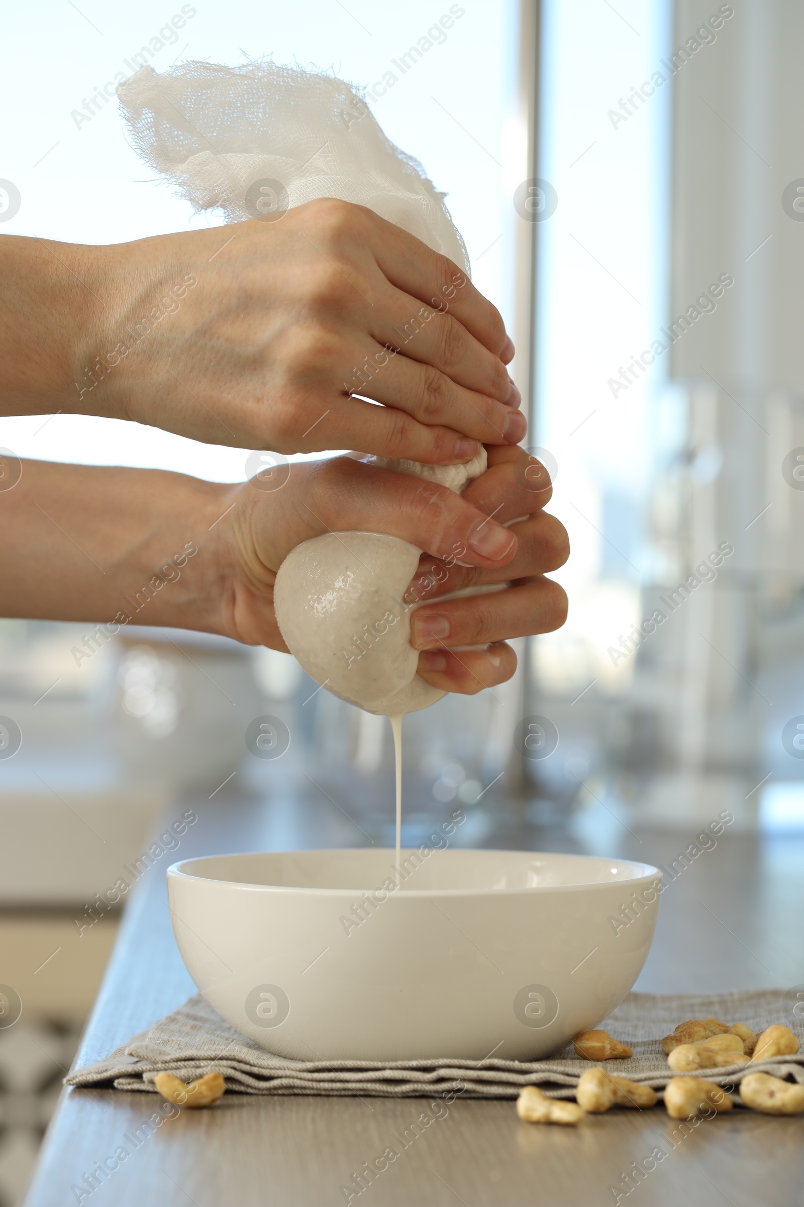 Photo of Woman straining cashew milk into bowl at wooden table with nuts indoors, closeup