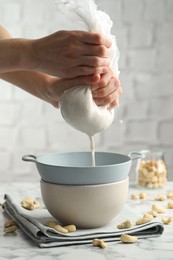 Woman straining cashew milk into colander with bowl at white marble table with nuts indoors, closeup