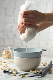 Photo of Woman straining cashew milk into colander with bowl at white marble table with nuts indoors, closeup