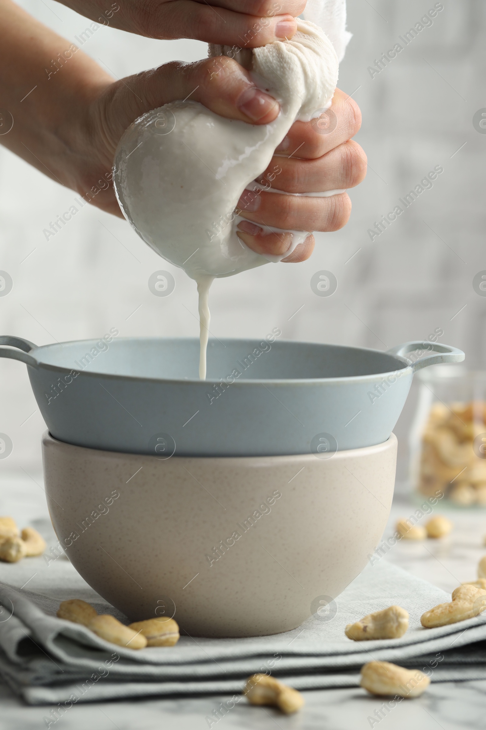 Photo of Woman straining cashew milk into colander with bowl at light table with nuts indoors, closeup