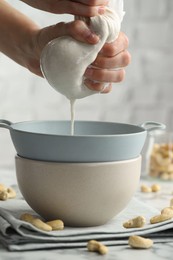 Woman straining cashew milk into colander with bowl at light table with nuts indoors, closeup