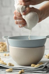 Photo of Woman straining cashew milk into colander with bowl at light table with nuts indoors, closeup