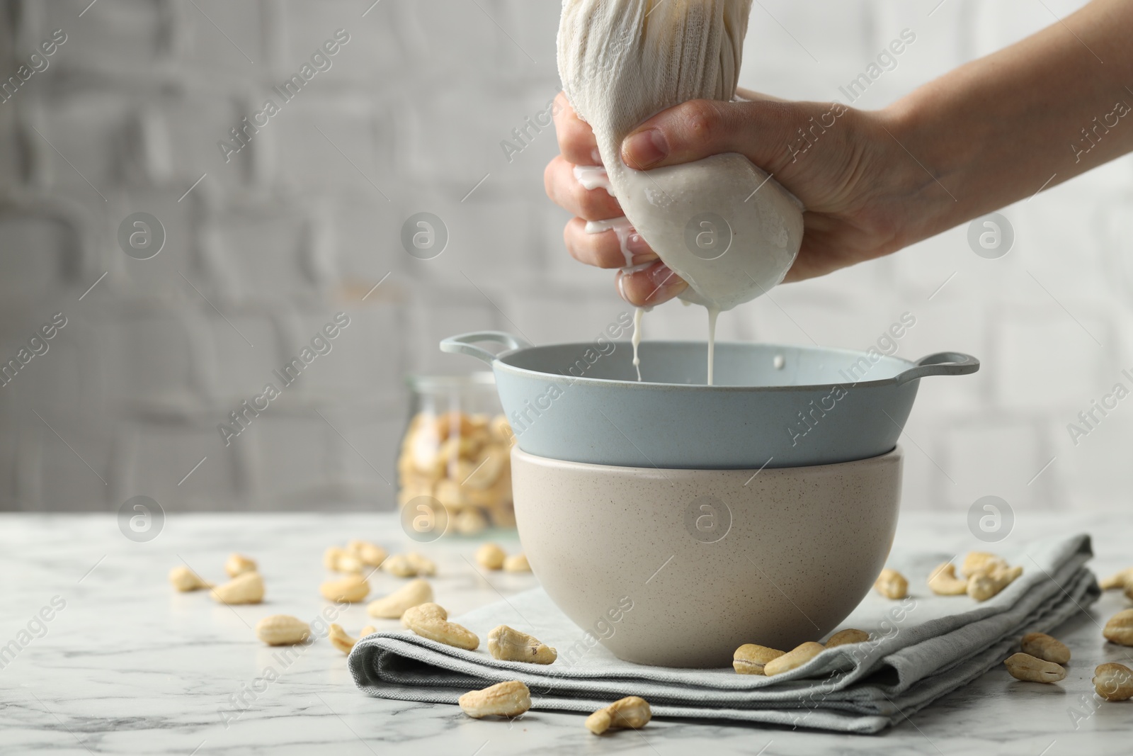 Photo of Woman straining cashew milk into colander with bowl at white marble table with nuts indoors, closeup. Space for text