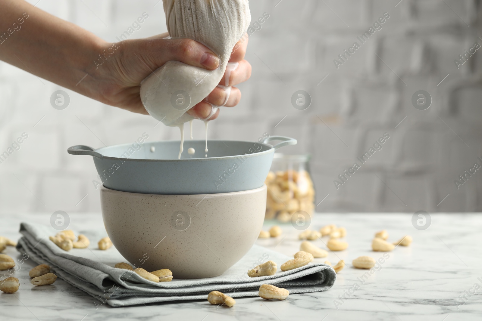 Photo of Woman straining cashew milk into colander with bowl at white marble table with nuts indoors, closeup. Space for text