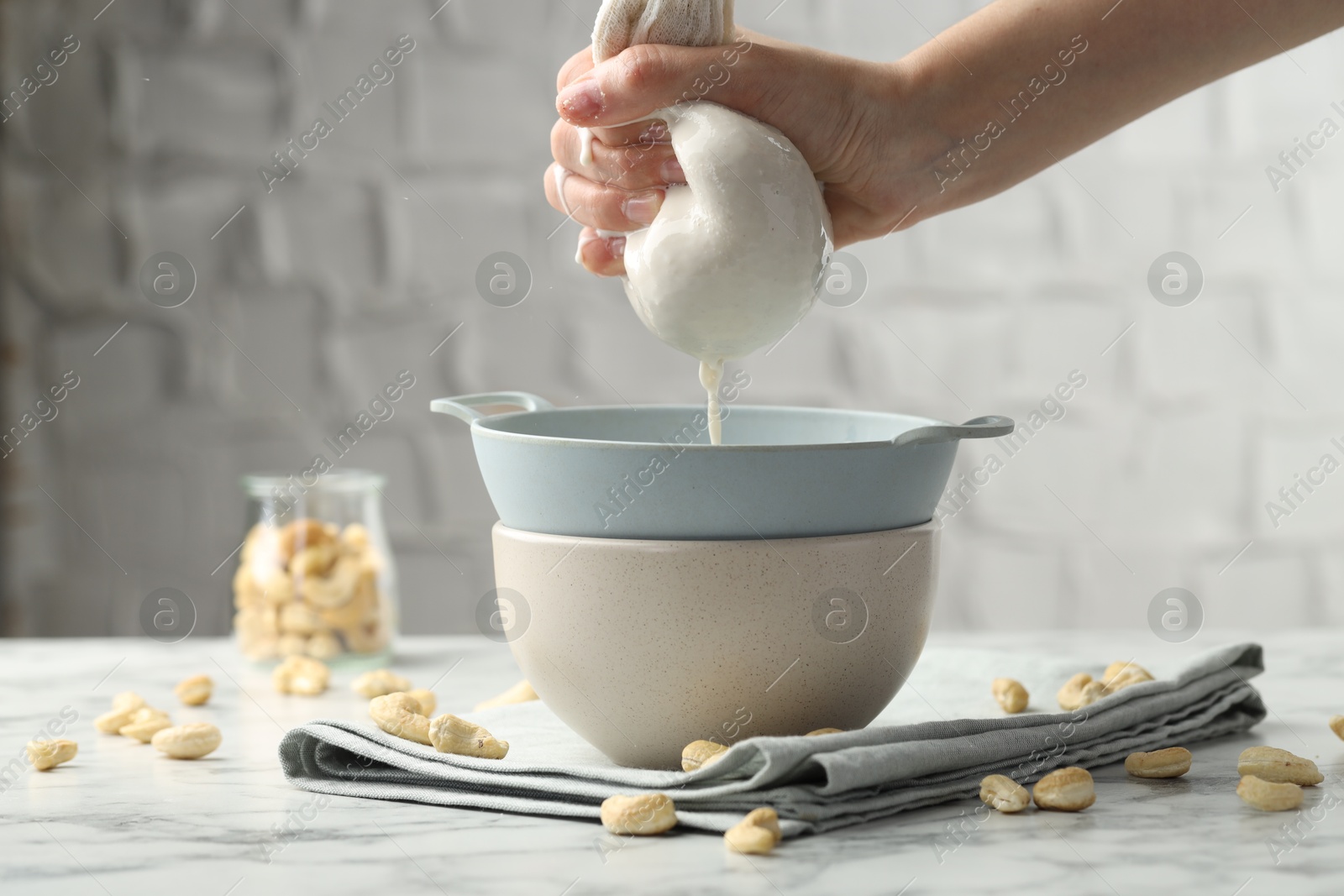 Photo of Woman straining cashew milk into colander with bowl at white marble table with nuts indoors, closeup