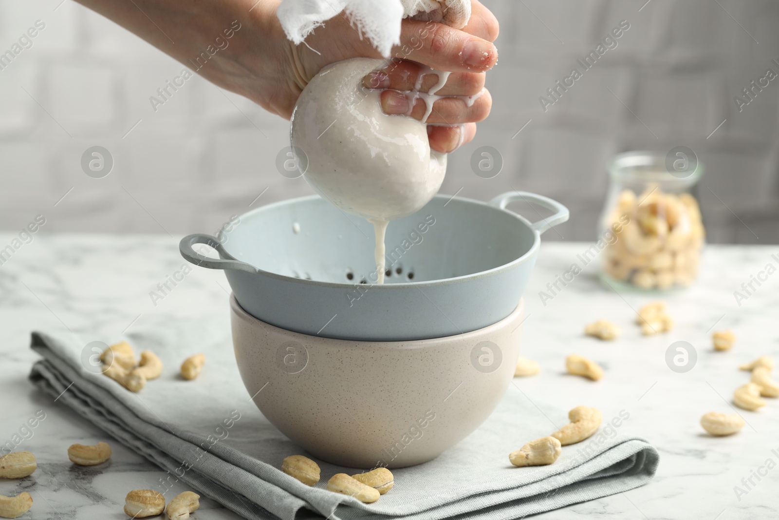 Photo of Woman straining cashew milk into colander with bowl at white marble table with nuts indoors, closeup