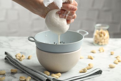 Photo of Woman straining cashew milk into colander with bowl at white marble table with nuts indoors, closeup