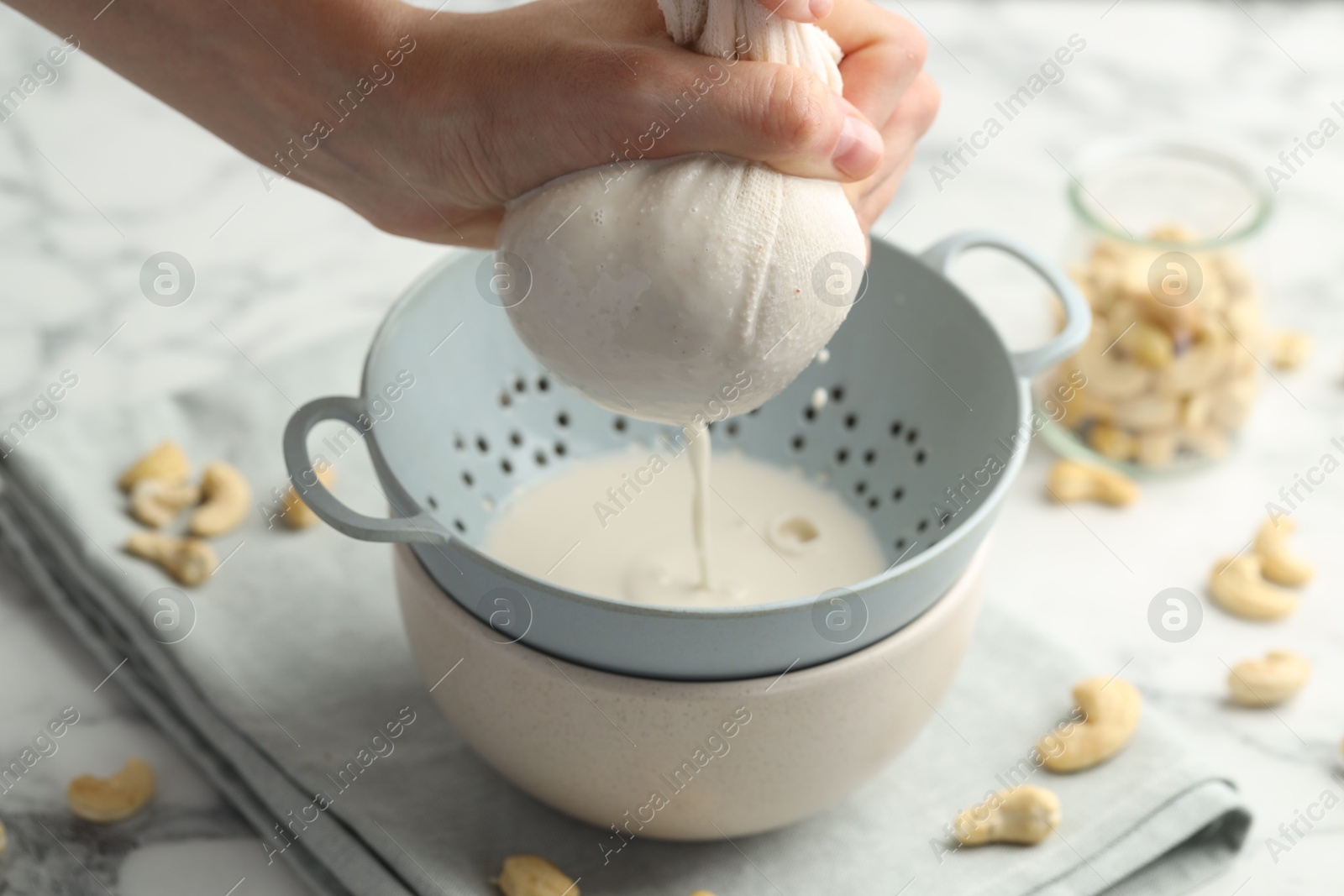 Photo of Woman straining cashew milk into colander with bowl at white marble table with nuts, closeup