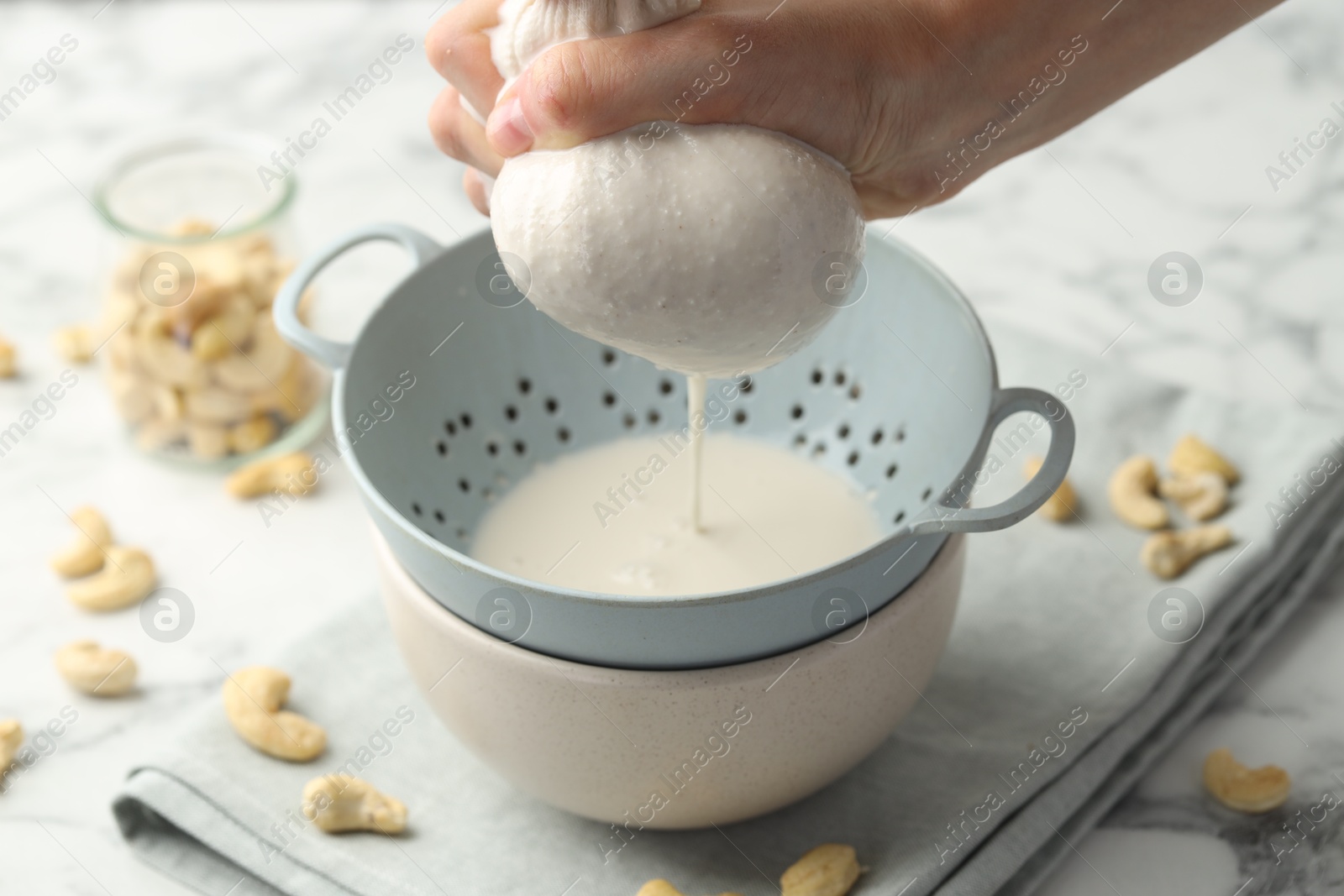 Photo of Woman straining cashew milk into colander with bowl at white marble table with nuts, closeup