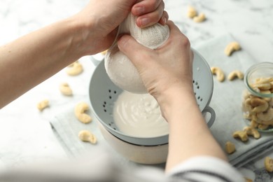 Woman straining cashew milk into colander with bowl at white table with nuts, closeup