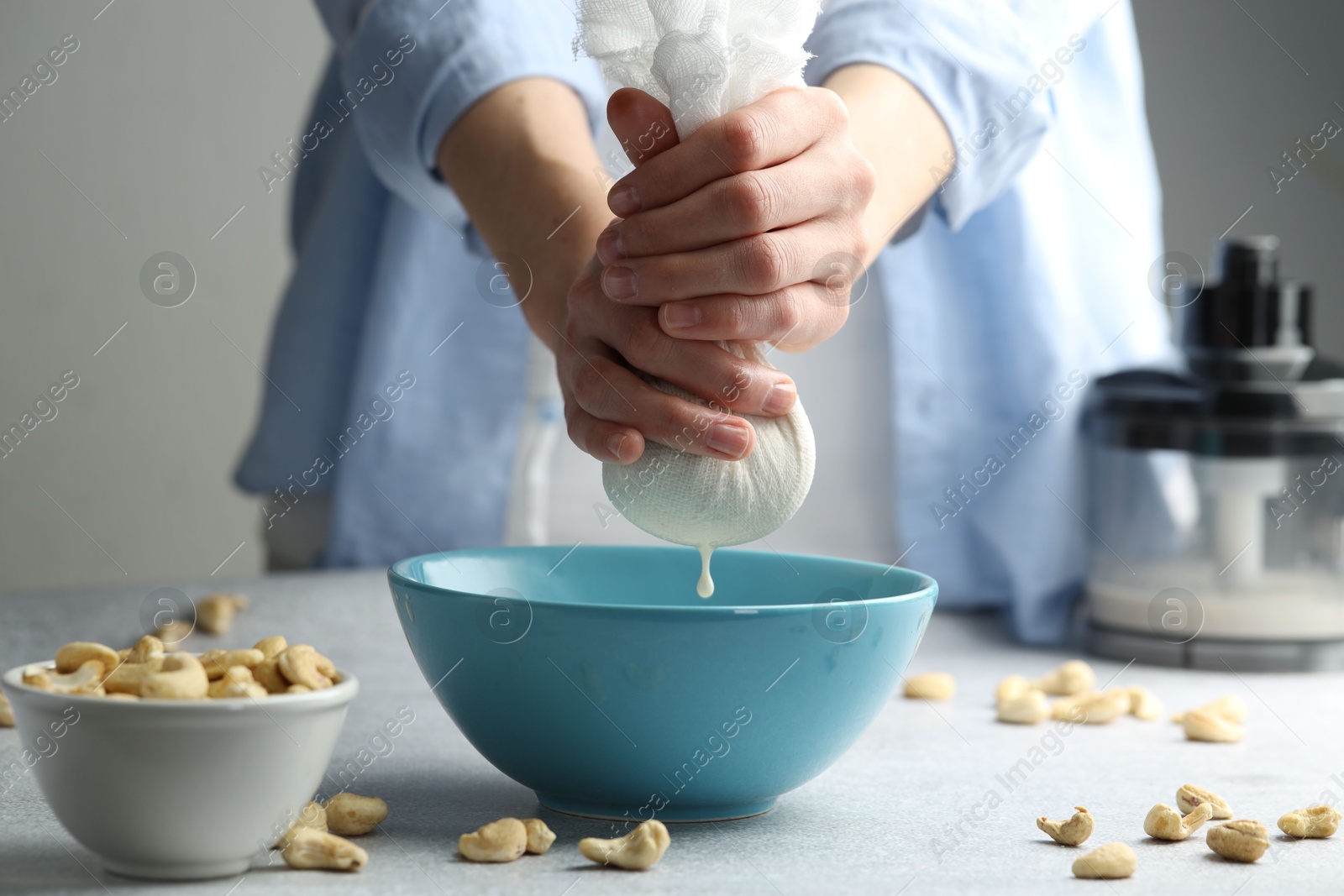 Photo of Woman straining cashew milk into bowl at light table with nuts indoors, closeup