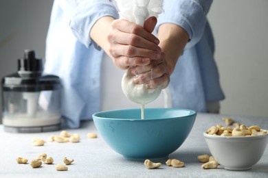 Photo of Woman straining cashew milk into bowl at light table with nuts indoors, closeup