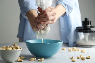 Woman straining cashew milk into bowl at light table with nuts indoors, closeup