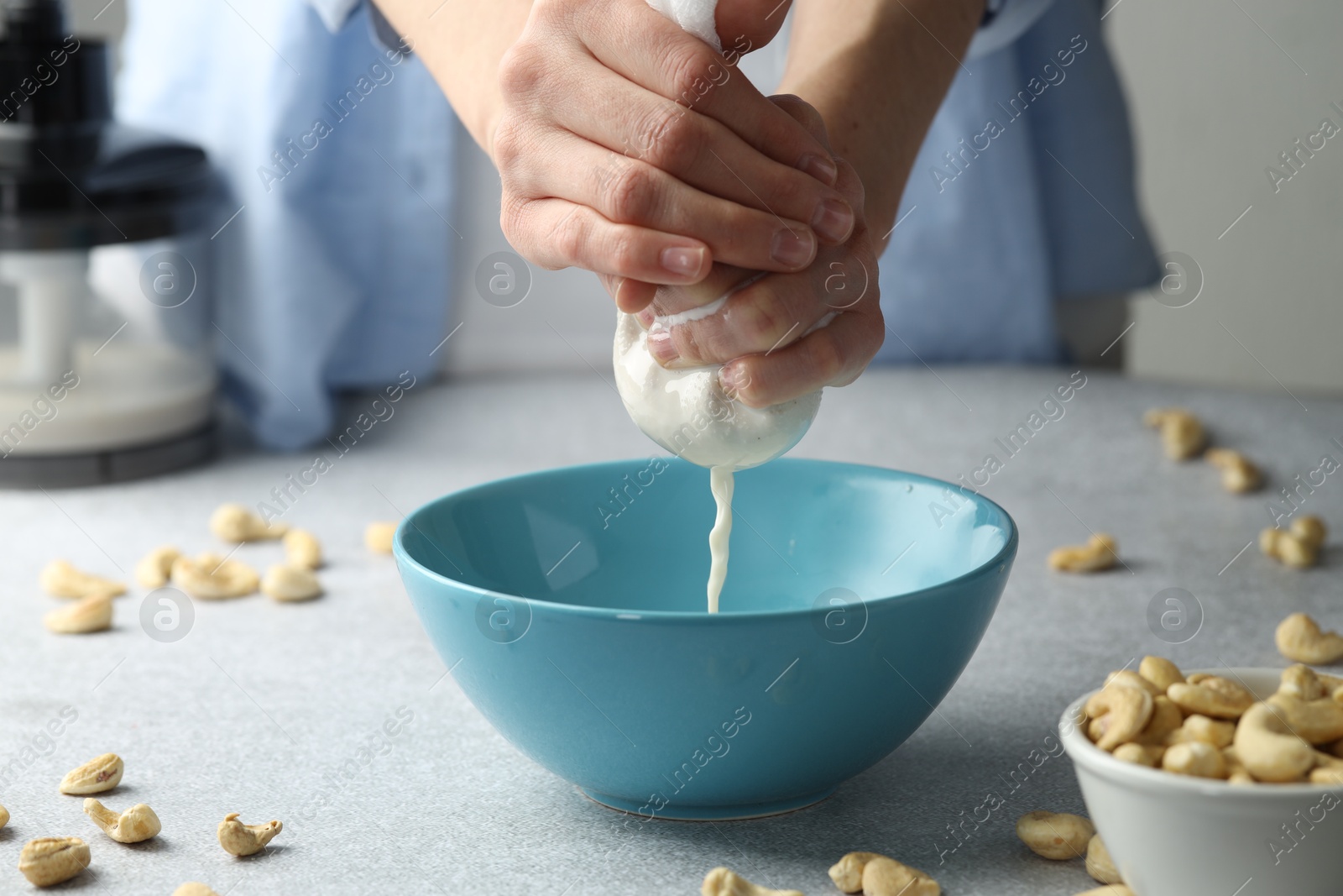 Photo of Woman straining cashew milk into bowl at light table with nuts indoors, closeup