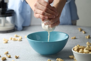 Woman straining cashew milk into bowl at light table with nuts indoors, closeup