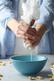 Photo of Woman straining cashew milk into bowl at light table with nuts, closeup