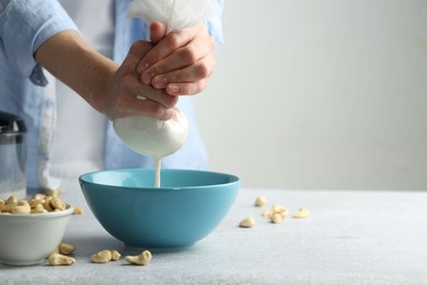 Woman straining cashew milk into bowl at light table with nuts indoors, closeup. Space for text