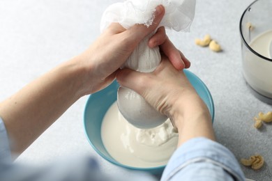 Photo of Woman straining cashew milk into bowl at light table with nuts, closeup