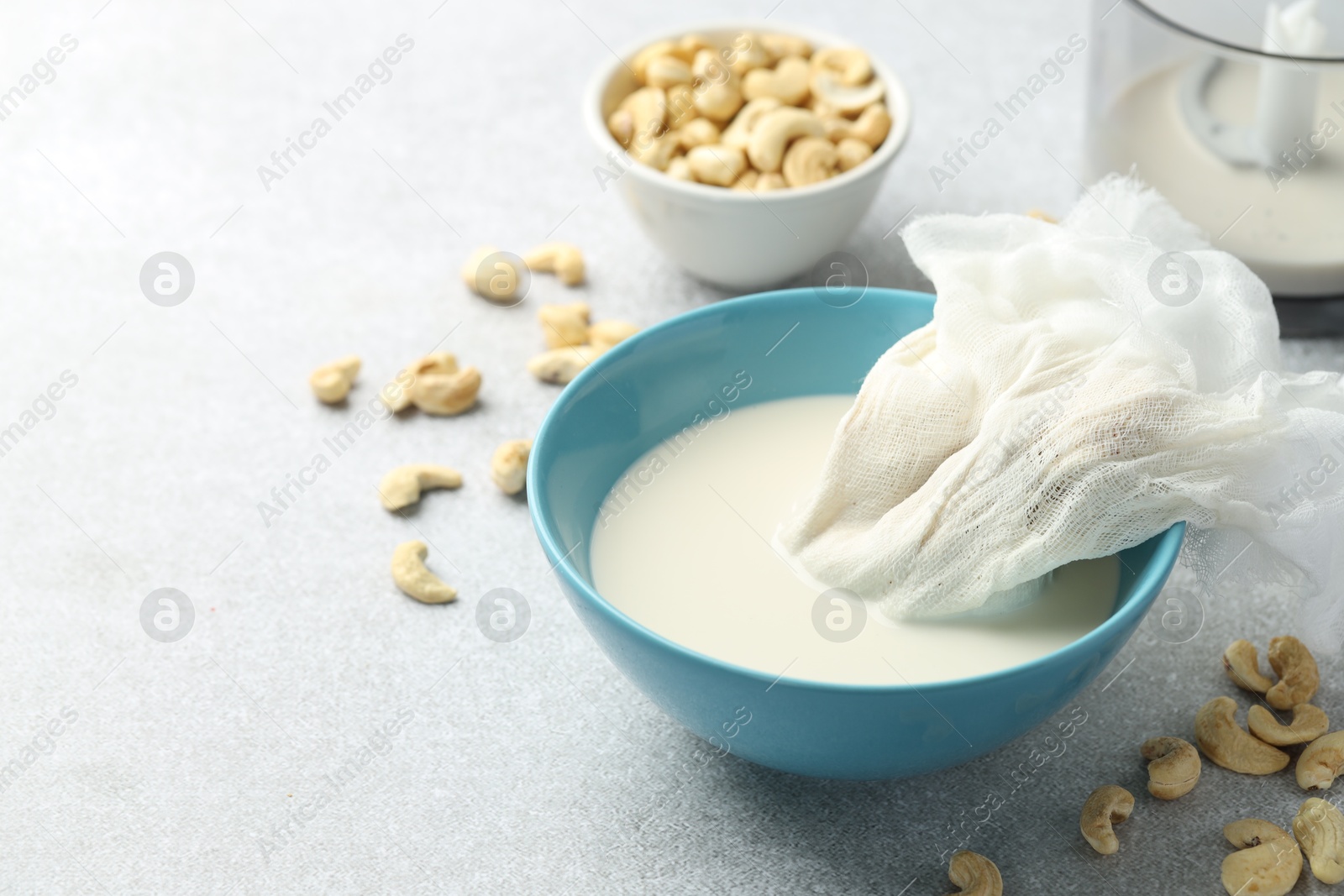 Photo of Making cashew milk. Cheesecloth and milk in bowl among nuts on grey table indoors, closeup. Space for text