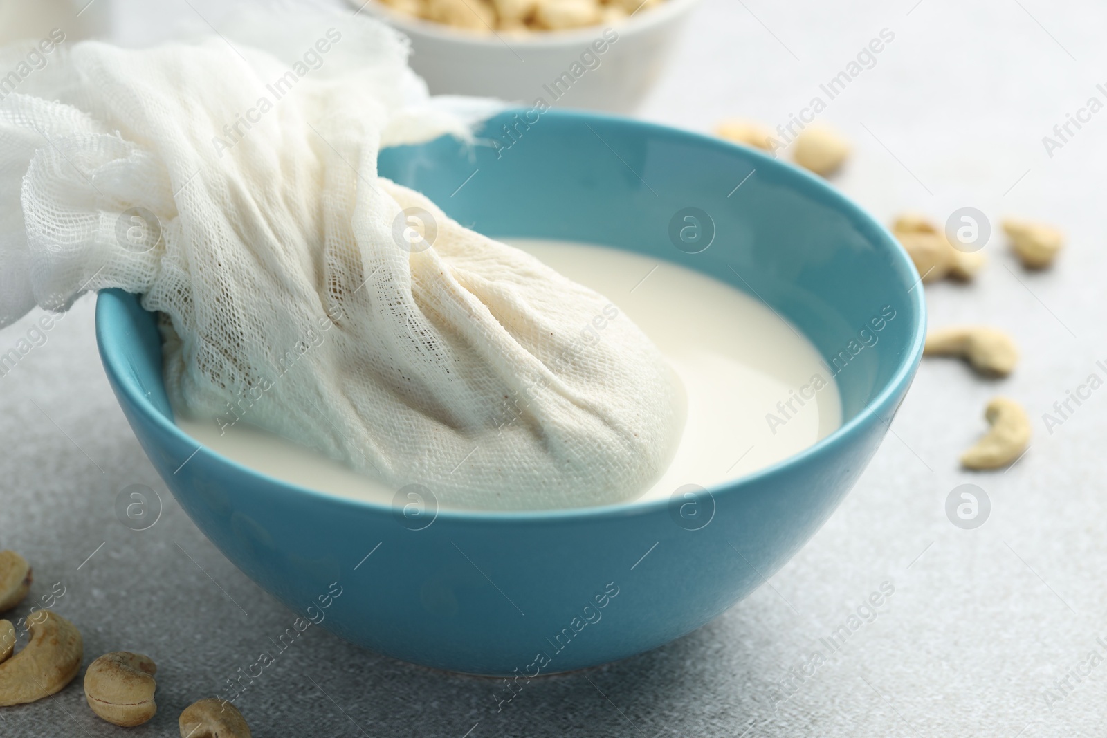 Photo of Making cashew milk. Cheesecloth and milk in bowl among nuts on grey table indoors, closeup