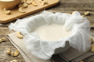 Photo of Making cashew milk. Cheesecloth and milk in bowl among nuts on wooden table, closeup