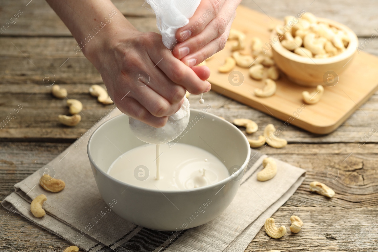 Photo of Woman straining cashew milk into bowl at wooden table with nuts, closeup