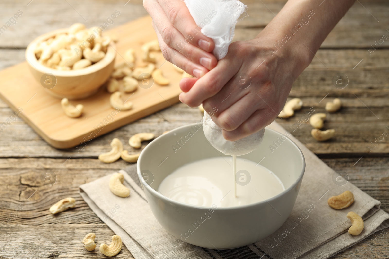 Photo of Woman straining cashew milk into bowl at wooden table with nuts, closeup