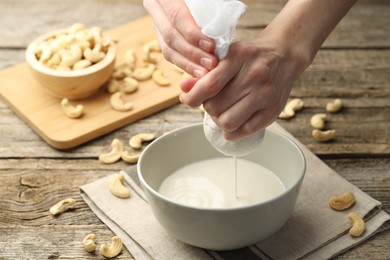 Woman straining cashew milk into bowl at wooden table with nuts, closeup