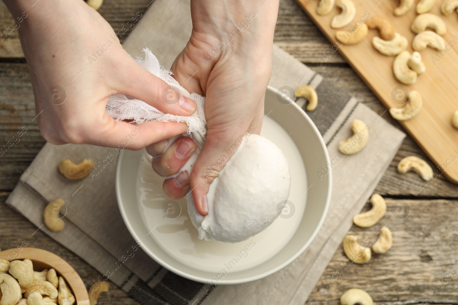 Photo of Woman straining cashew milk into bowl at wooden table with nuts, top view