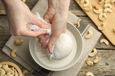 Woman straining cashew milk into bowl at wooden table with nuts, top view