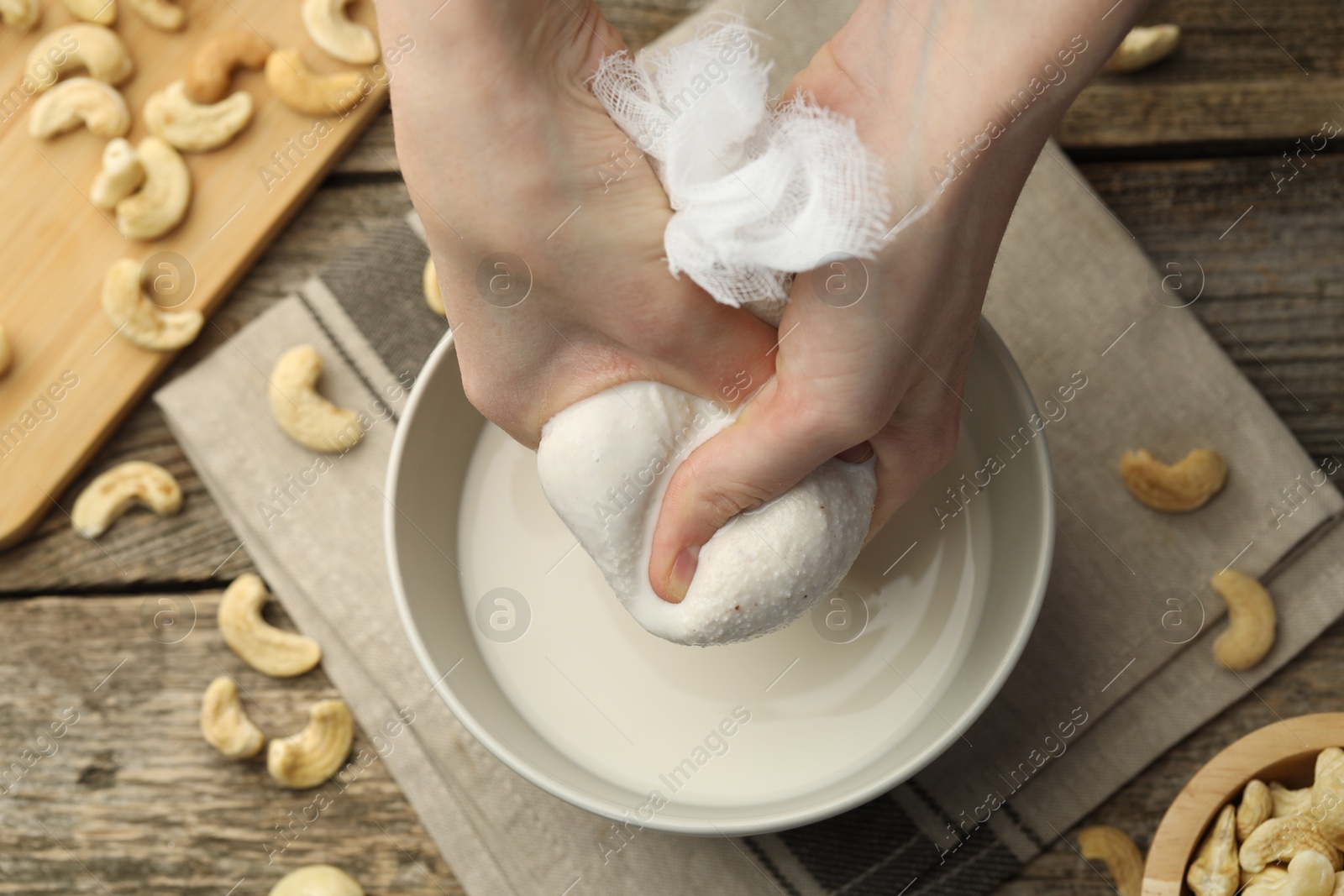 Photo of Woman straining cashew milk into bowl at wooden table with nuts, top view