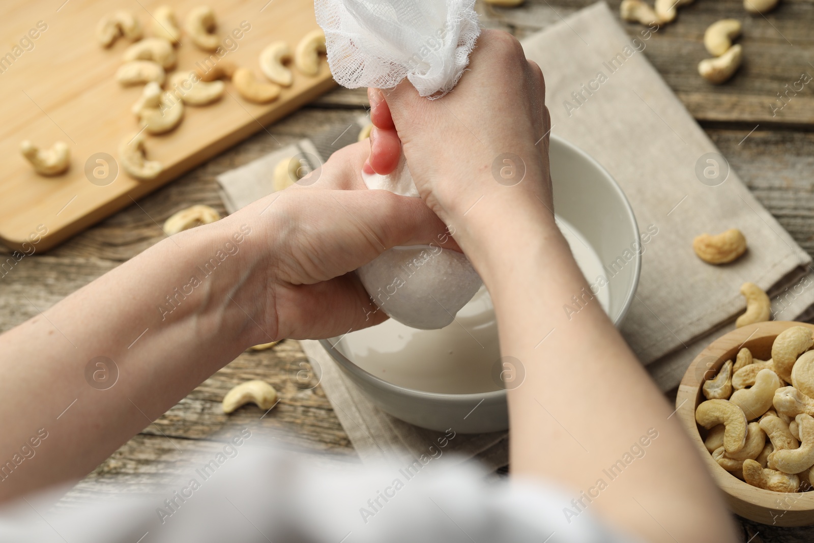 Photo of Woman straining cashew milk into bowl at wooden table with nuts, closeup